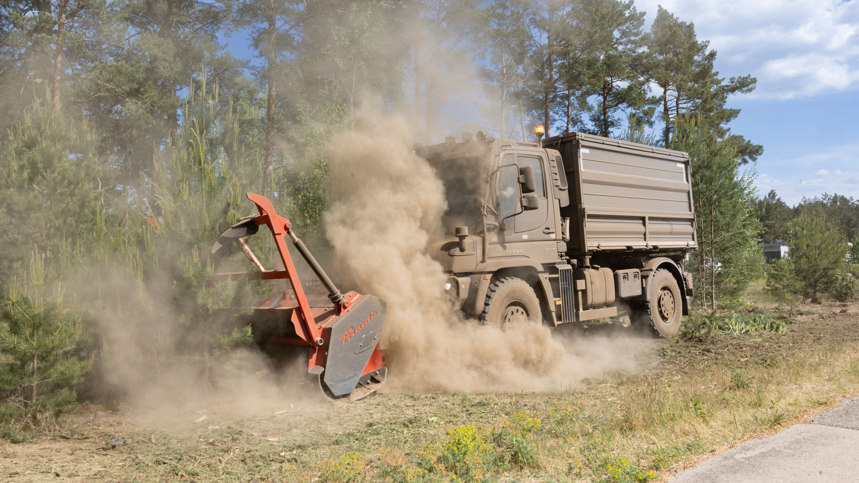 Un camion Mercedes‑Benz équipé d'un broyeur attelé travaille dans une zone forestière et soulève un nuage de poussière en traitant la végétation.