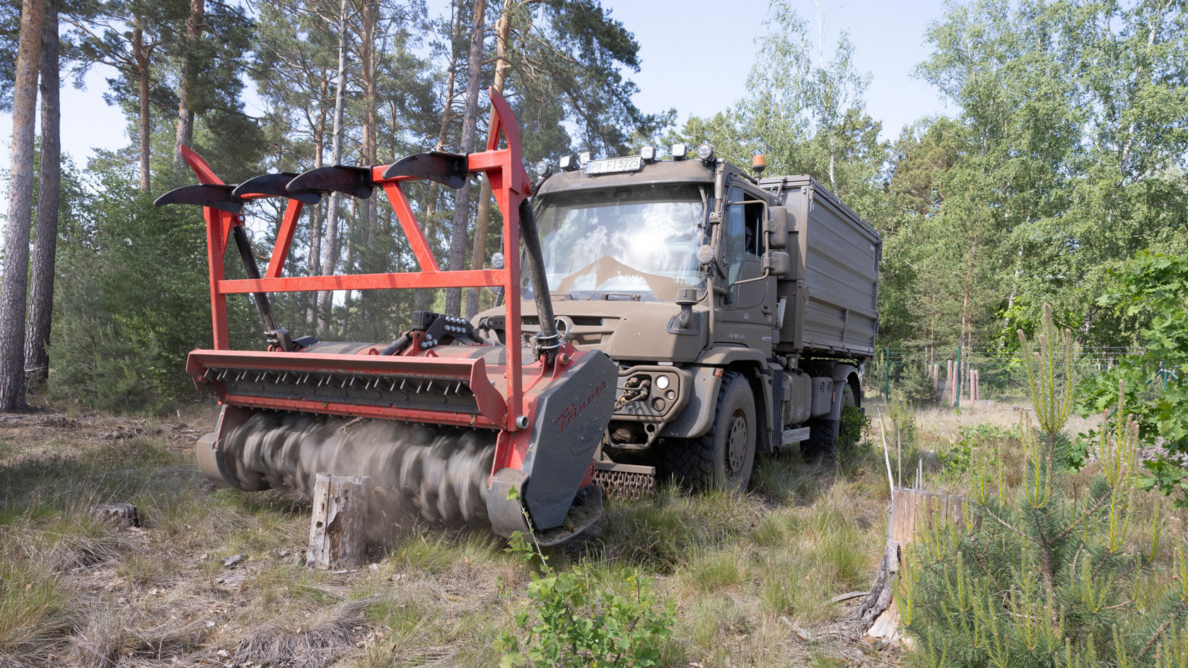 Un camion Mercedes-Benz équipé d'une machine spéciale de travail du sol travaille dans une zone forestière, tout en ameublissant le sol et en soulevant de la terre.