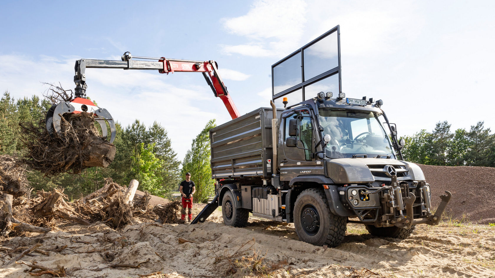Un véhicule de travail Mercedes‑Benz soulève une grande charge de bois à l'aide d'une grue. À côté du véhicule se tient un ouvrier en tenue de travail.