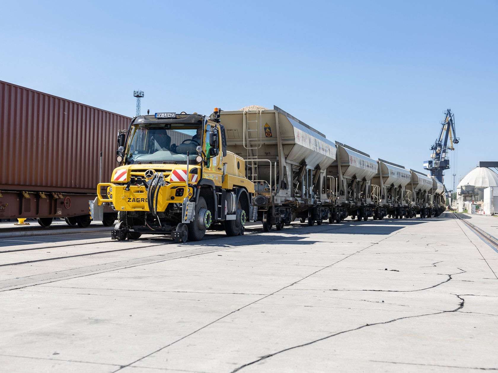 Ein Mercedes-Benz Unimog zieht einen langen Zug von Containern und Kippern in einem Hafenbereich, umgeben von Krane und Ladeinfrastruktur.