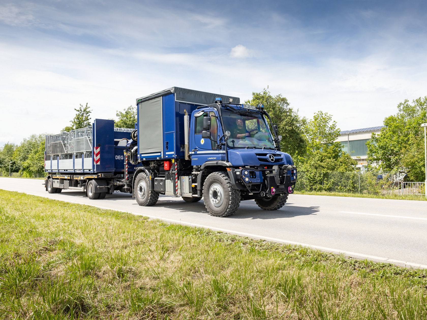 Ein blauer Mercedes-Benz Unimog fährt auf einer Straße und zieht einen Anhänger mit einer großen Ladefläche.