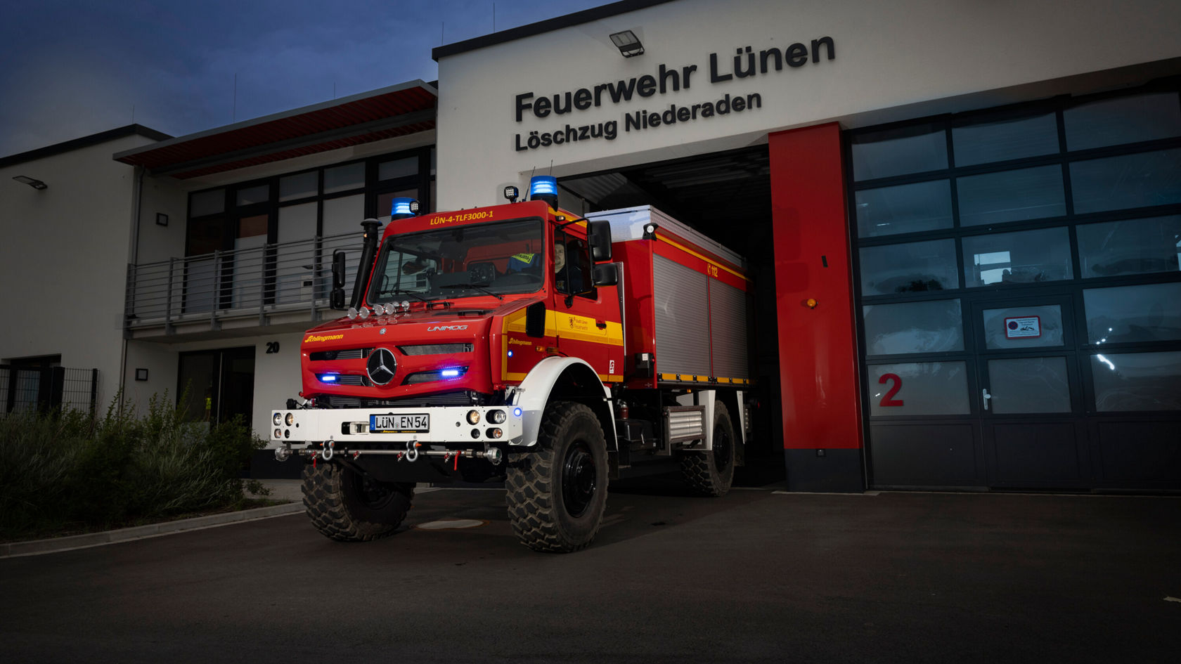 Un véhicule de pompiers Mercedes‑Benz est stationné devant la caserne de pompiers de Lünen‑Niederaden. Le véhicule est équipé de gyrophares et prêt pour la lutte contre les incendies.