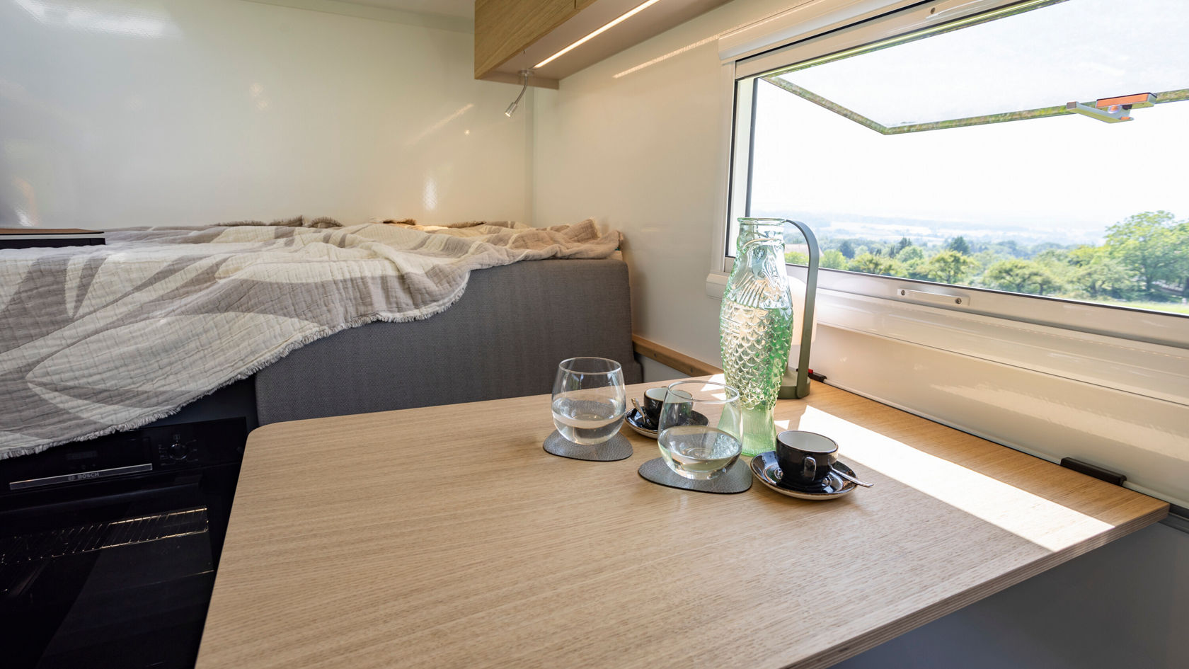 Un intérieur douillet avec un lit à l'arrière-plan et une table au premier plan. Une grande fenêtre donne sur un paysage verdoyant à l'extérieur.