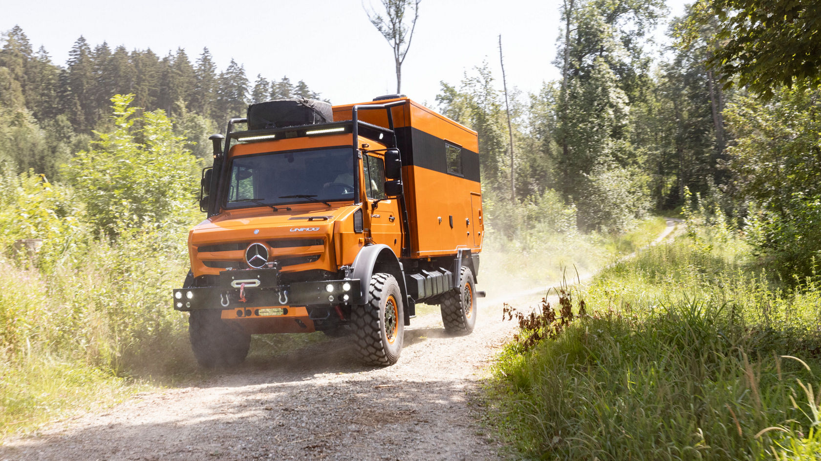 Un camping-car Unimog de couleur orange circule sur un chemin forestier poussiéreux. Le camping-car a de gros pneus et est conçu pour des sorties tout-terrain.