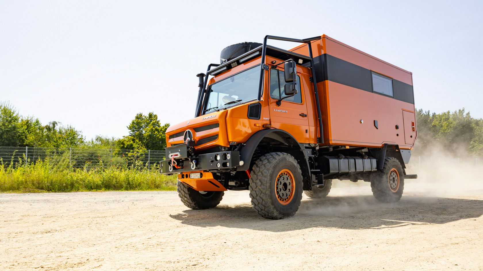 Un camping-car Unimog orange roule sur un sol sableux. Il est équipé de grands pneus et d'un arceau de sécurité.