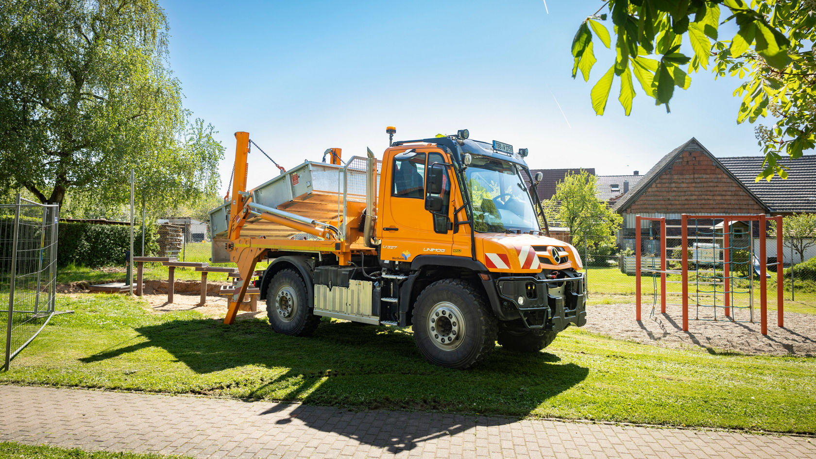 Een oranje Unimog staat op een grasveld in een speeltuin. De Unimog heeft een grote laadbak en is omgeven door bomen en een speeltoestel.