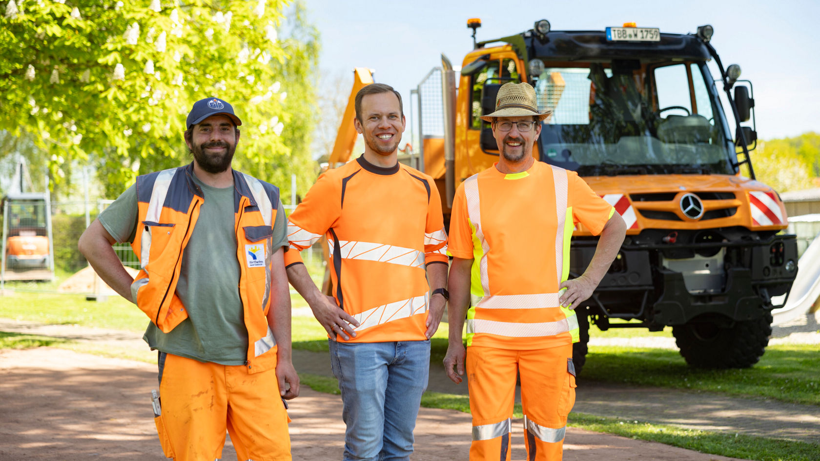 Drie mannen in oranje werkkleding staan glimlachend in een speeltuin. Op de achtergrond is een Unimog te zien.