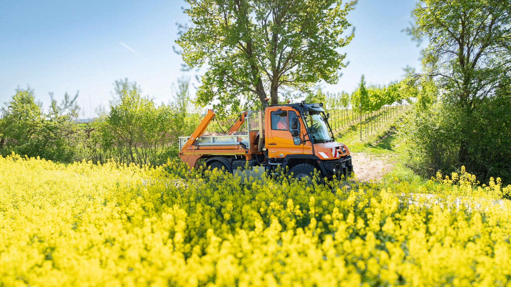 Een oranje Unimog rijdt op een weg tussen bloeiende koolzaadvelden en bomen.