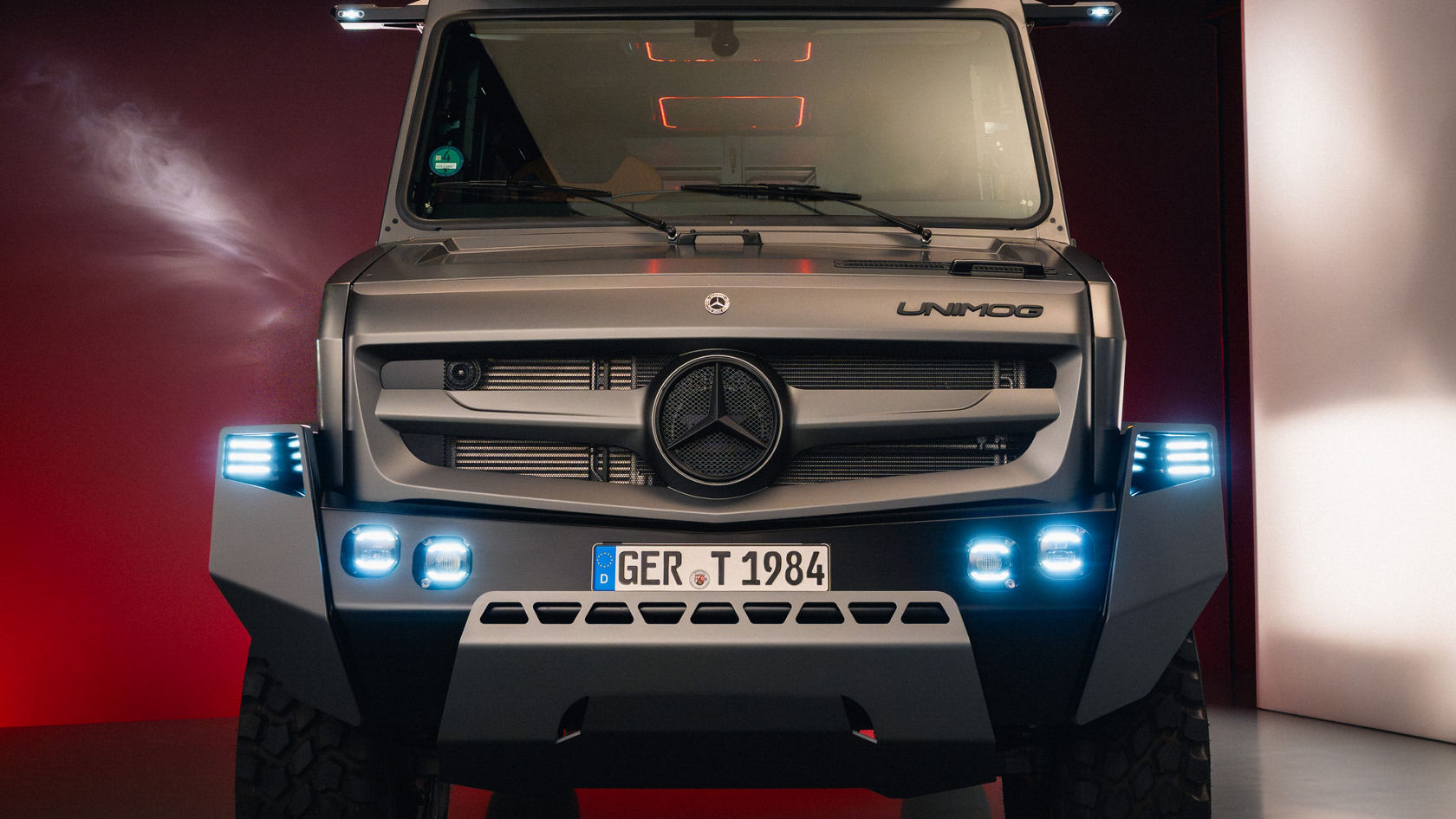A Mercedes-Benz Unimog SUV with a robust design and high ground clearance, set against a red background, is distinguished by its striking headlights.
