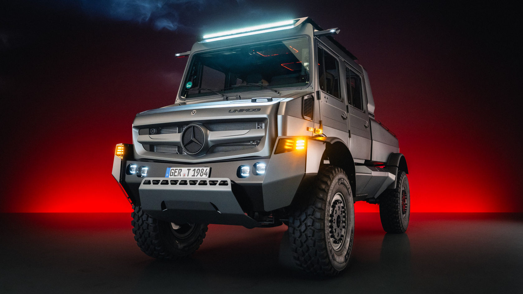 A Mercedes-Benz Unimog stands in dramatic lighting, with the headlights and an LED light on the roof illuminated. The off-roader has large tires and a striking, robust build.