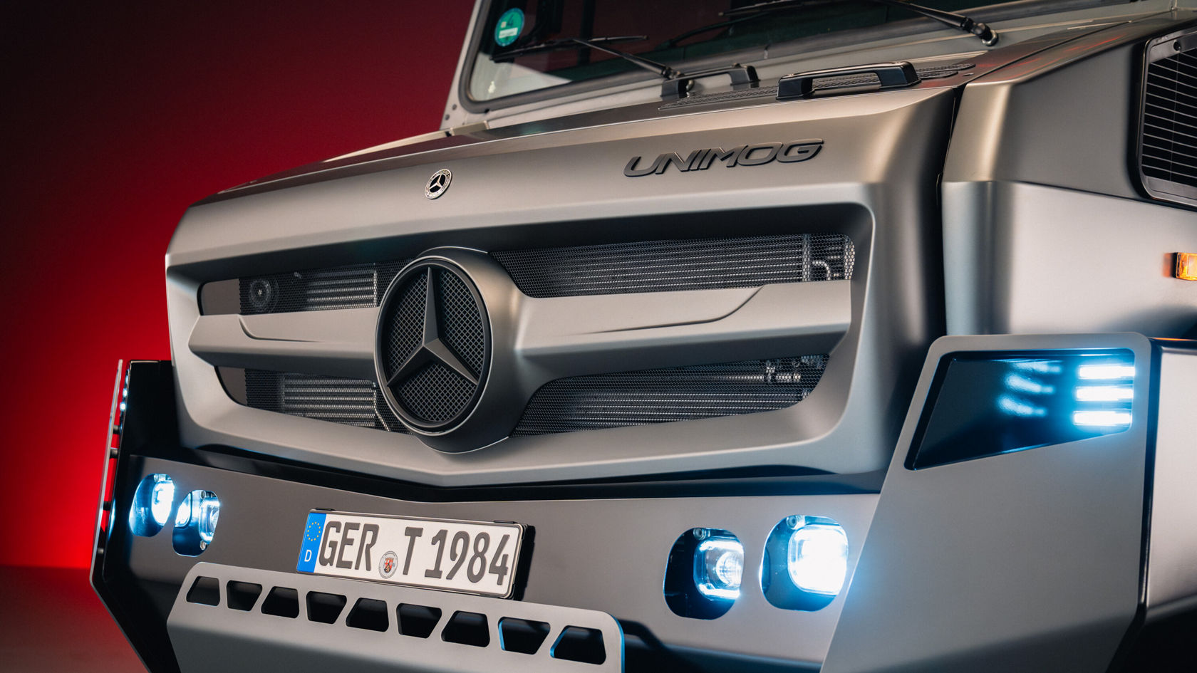 Ein Unimog von Mercedes-Benz steht beleuchtet vor einem roten Hintergrund, umgeben von leichtem Nebel. Der robuste Truck zeigt aggressive Designelemente und ist mit starken Reifen ausgestattet.