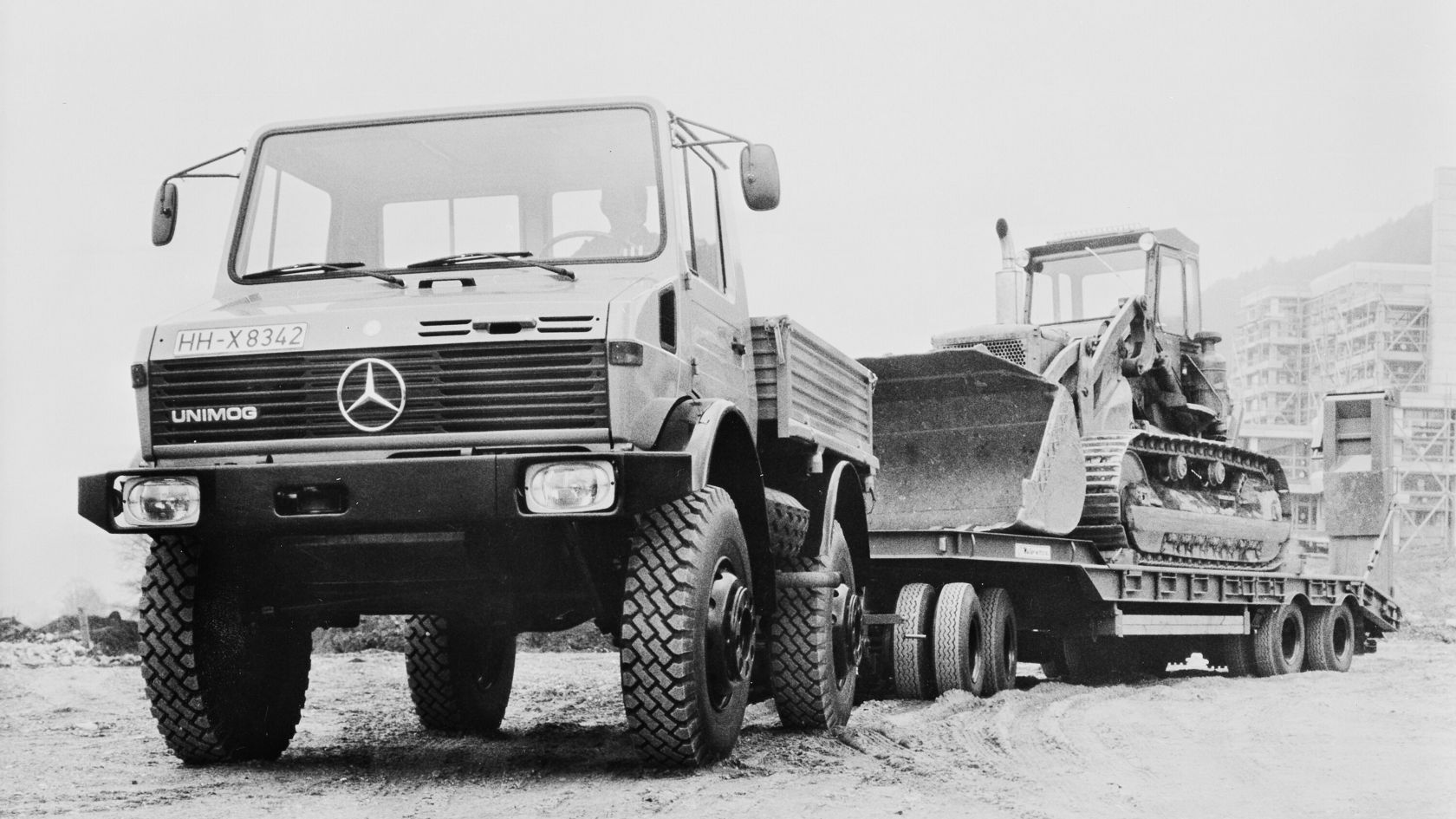 A Mercedes-Benz Unimog is on a construction site, while an excavator on a trailer is visible in the background. The scene shows heavy work vehicles in an industrial environment.