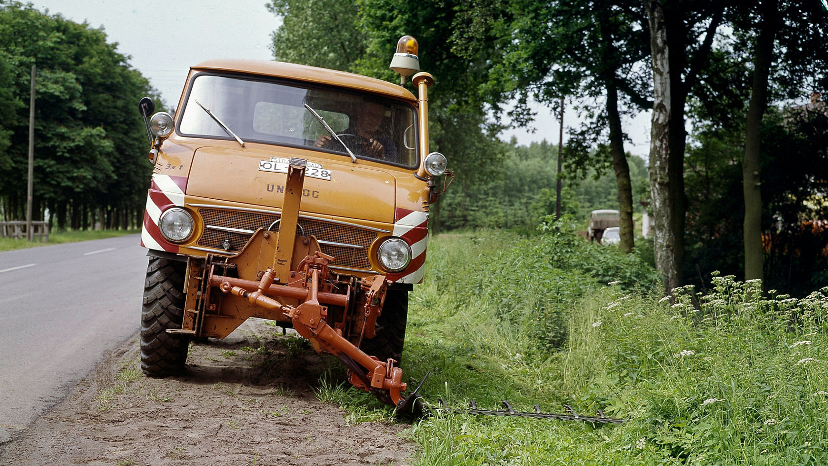 Een oranje Mercedes-Benz Unimog staat aan de kant van de weg. Hij heeft een gele waarschuwingslamp en een werktuig aan de voorzijde, terwijl bomen en een weide op de achtergrond zichtbaar zijn.