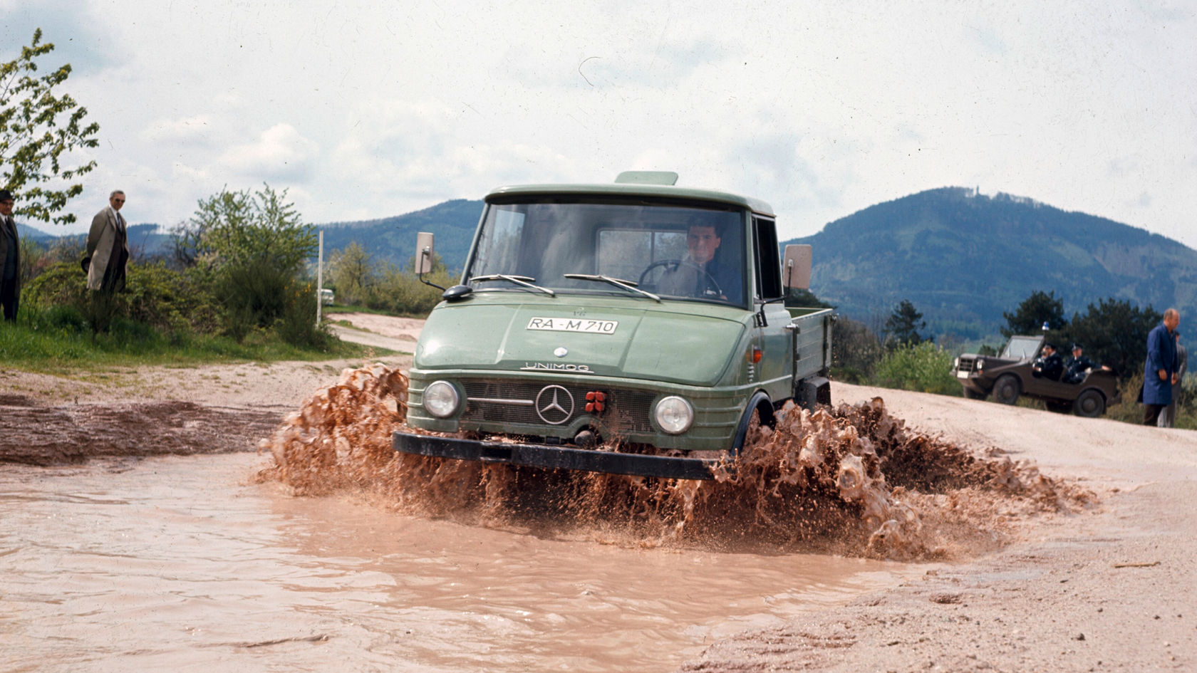 A Mercedes-Benz Unimog drives through a muddy puddle, splashing water and mud. People and rolling hills are visible in the background.
