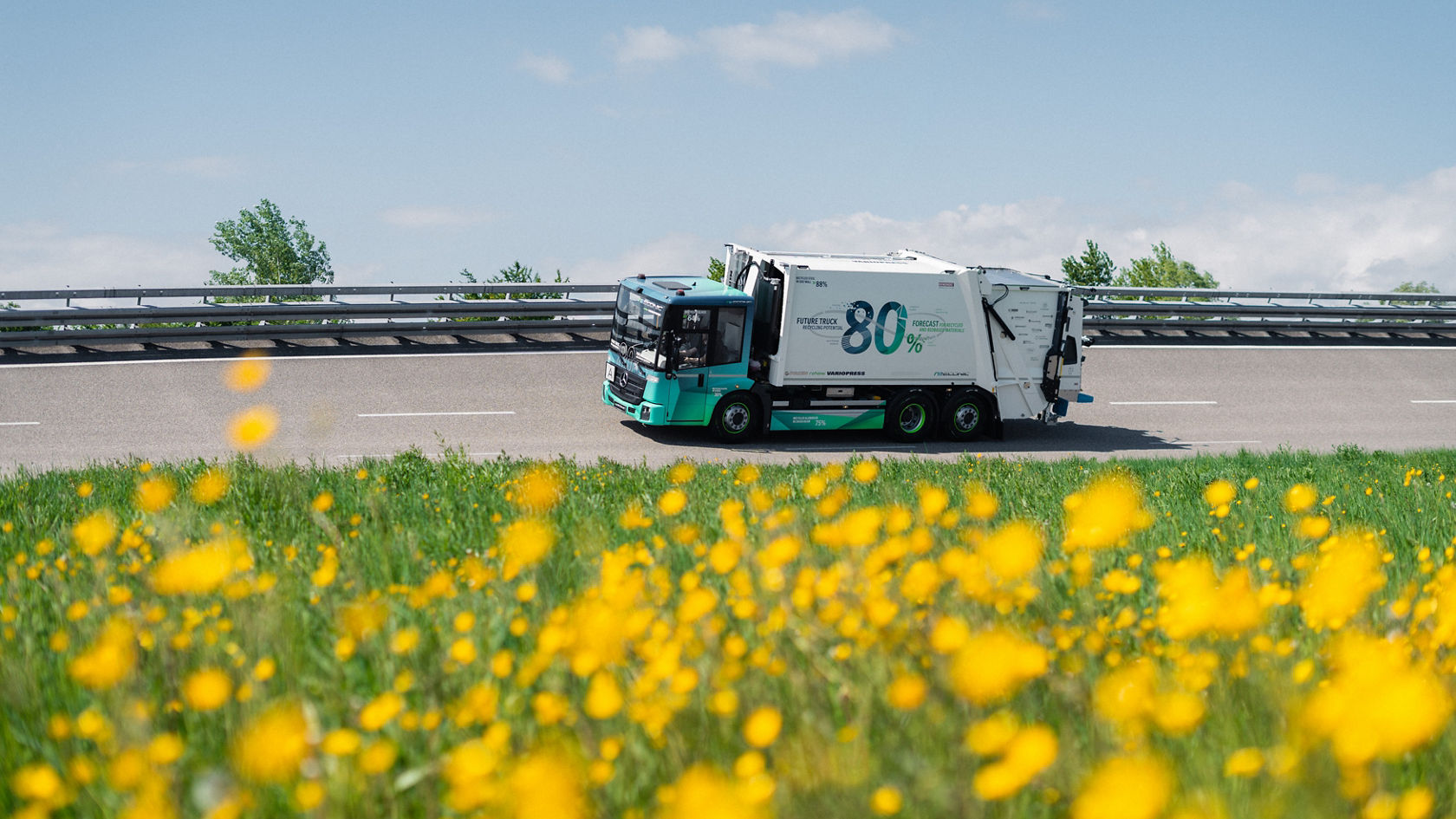 Mercedes-Benz eEconic Future Truck fährt hinter einer blühenden Löwenzahnwiese auf der Straße.