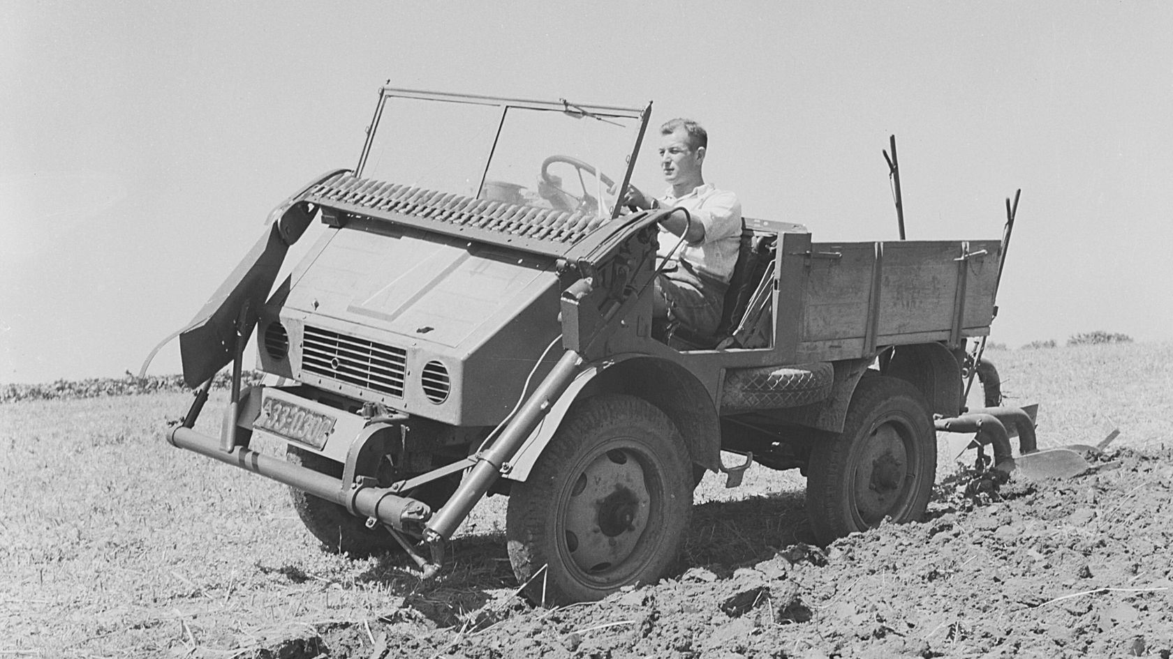 Ein Mercedes-Benz Unimog fährt über ein Feld, während ein Fahrer am Steuer sitzt und das Fahrzeug lenkt. Das Gelände ist uneben und bearbeitet.
