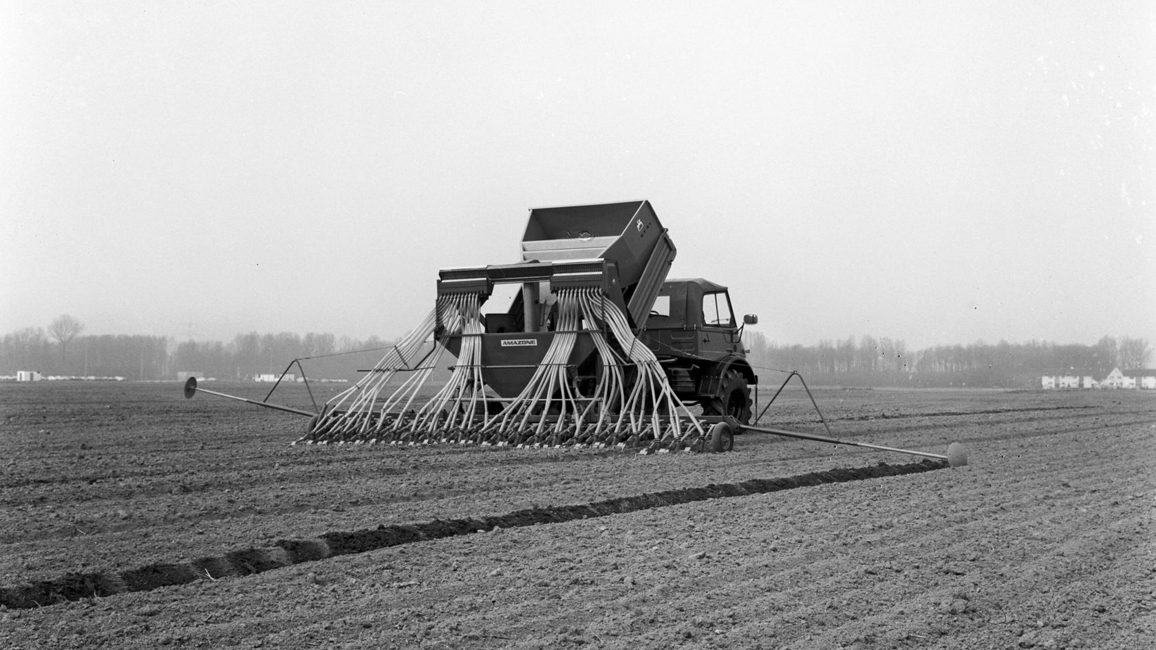 A Mercedes-Benz Unimog is standing in a field and is being used to apply seed. The machine has a seeding bar with several hoses.