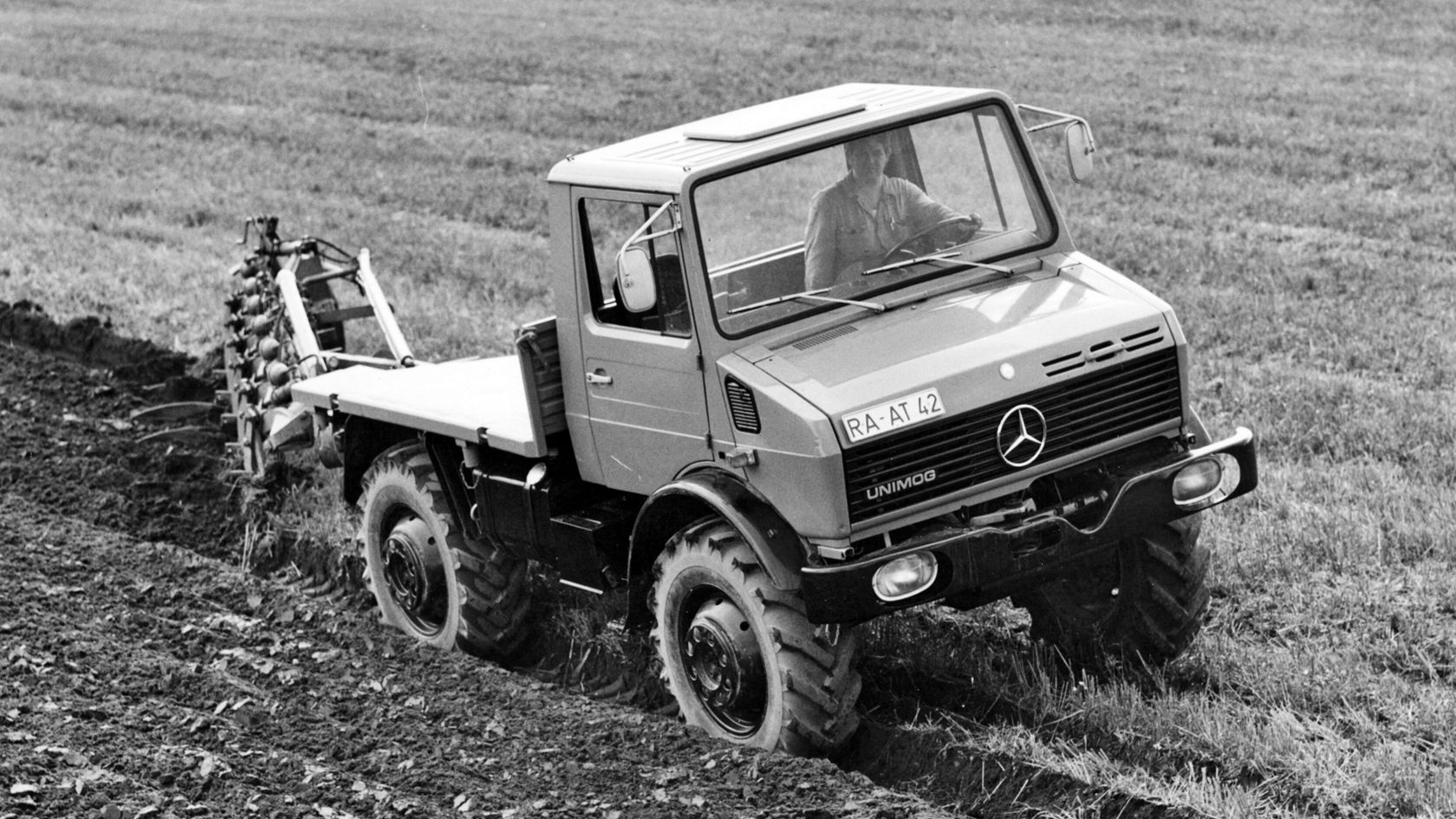 A Mercedes-Benz Unimog drives across a field and plows the soil. The driver is visible and is operating the vehicle, which is used for agricultural work.
