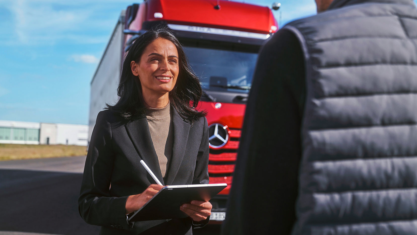 Eine Frau in einem schwarzen Blazer spricht mit einem Mann. Sie hält ein Tablet in der Hand und lächelt ihn an. Im Hintergrund steht ein roter Mercedes-Benz Lkw.
