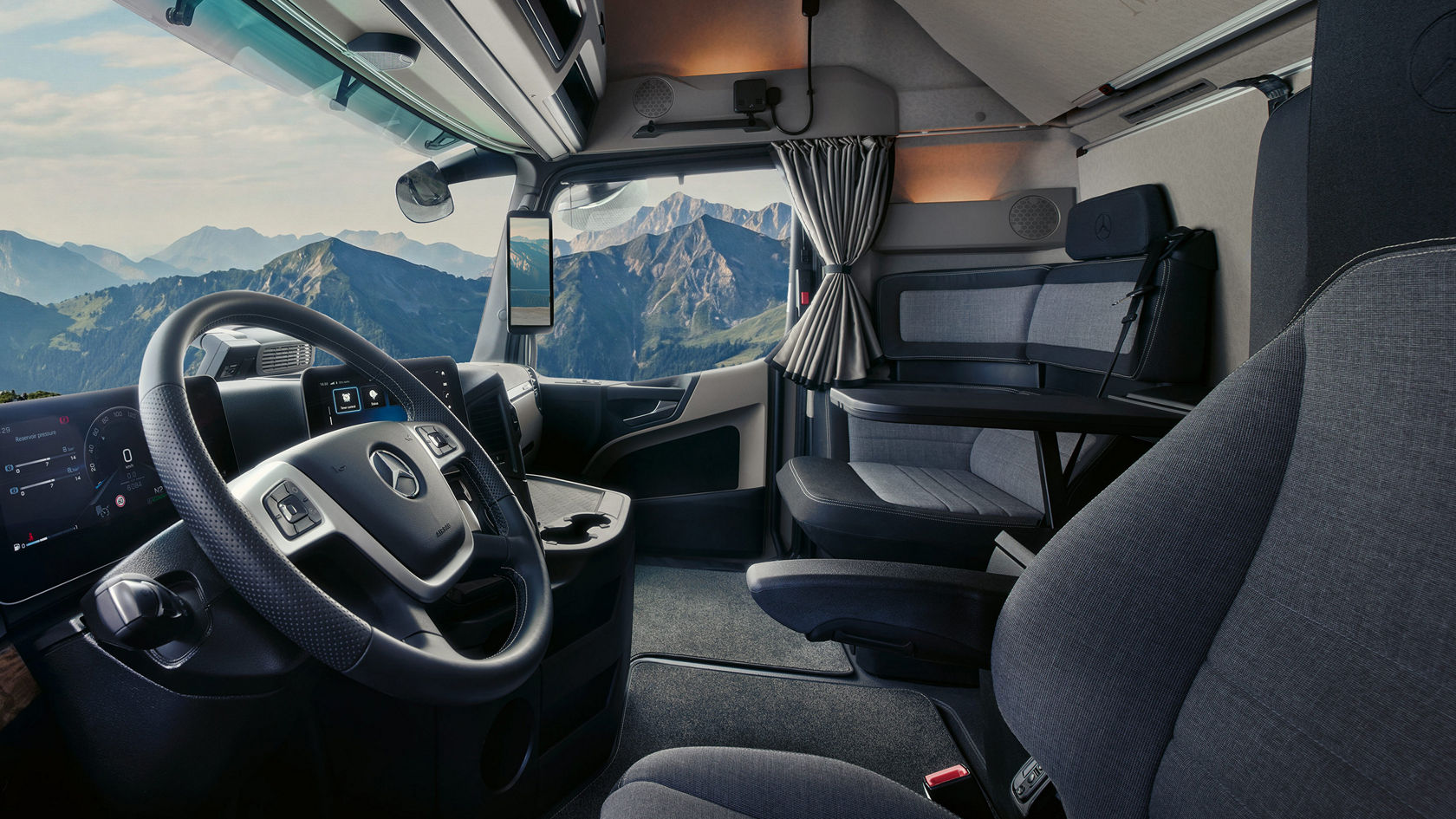 Das Innere der Fahrerkabine eines LKW. Man blickt auf das moderne Cockpit mit Lenkrad, digitalen Bildschirmen und der großen Frontscheibe. Im Hintergrund ist eine schöne Alpenlandschaft.