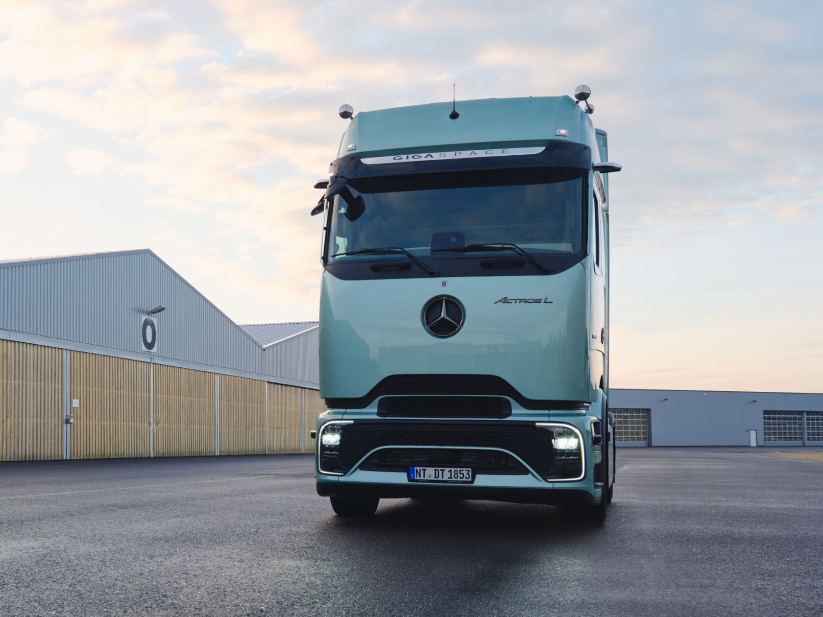 Frontansicht eines hellgrünen Mercedes-Benz Actros L Lkw, der auf nassem Asphalt vor einer Reihe von Industriehallen geparkt ist.