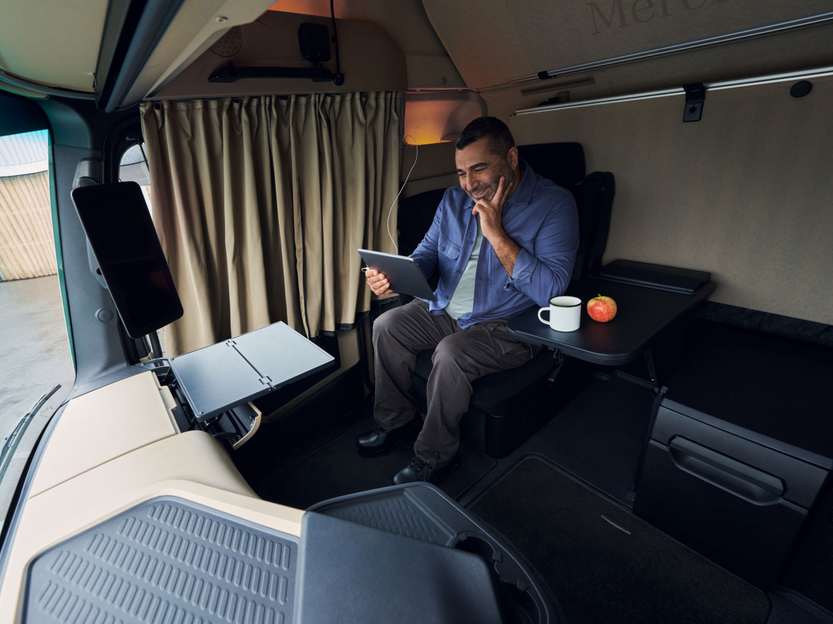 Ein Mann sitzt entspannt im geräumigen Innenraum einer Lkw-Fahrerkabine und schaut auf ein Tablet. Auf einem kleinen Tisch neben ihm stehen eine Tasse und ein Apfel.