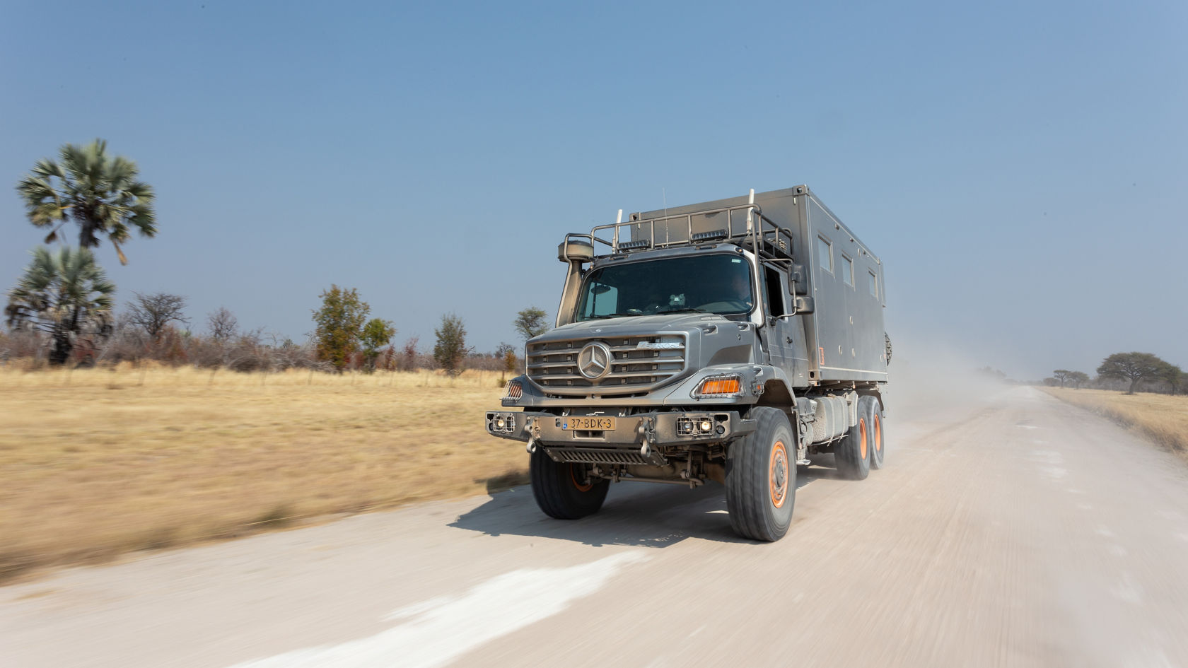 Ein Mercedes-Benz Zetros fährt auf einer staubigen Straße durch eine offene Landschaft mit vereinzelten Palmen und trockenem Gras.