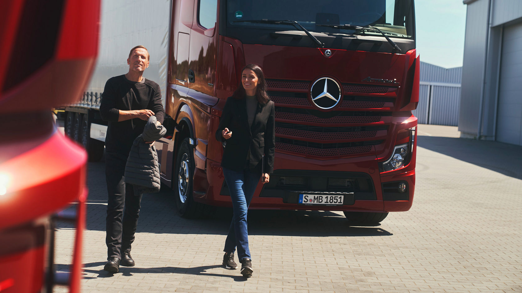 Ein Mann und eine Frau gehen auf einem gepflasterten Hof. Im Hintergrund steht die Front eines großen, roten Mercedes-Benz Lkw.