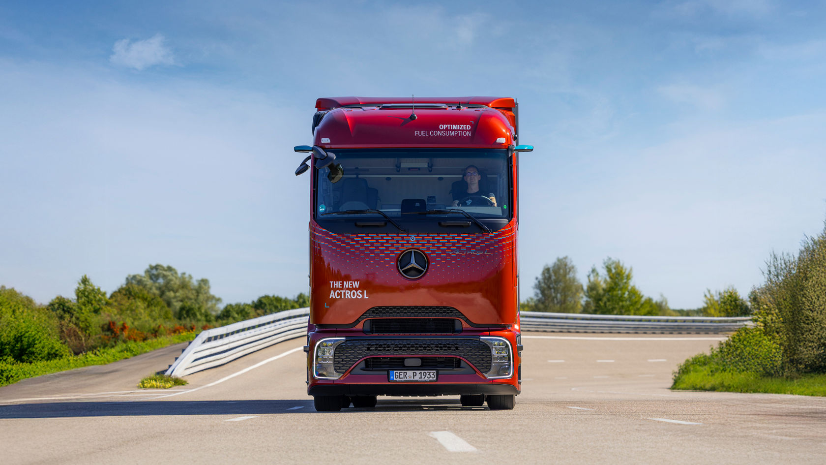 Ein roter Actros L fährt in Richtung der Kamera auf einer breiten, leeren Straße mit grünen Büschen links und rechts.