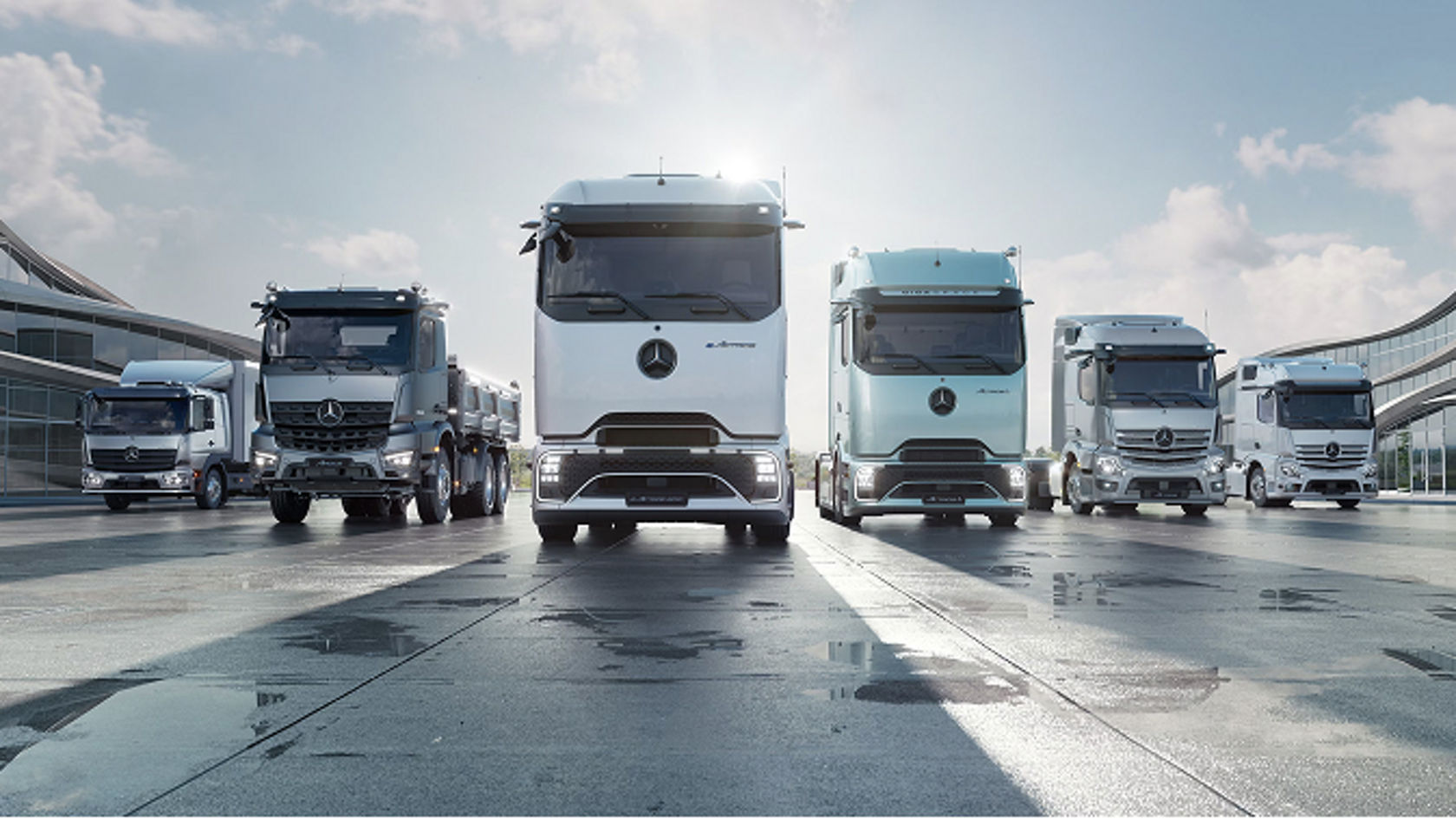 Das Bild zeigt eine Reihe von fünf Mrecedes-Benz Trucks, die auf einer leicht feuchten, asphaltierten Fläche vor einem futuristisch anmutenden Gebäude stehen. Die Lastwagen sind in verschiedenen Grautönen und einem hellen Türkis gehalten, wobei die beiden mittleren Fahrzeuge am größten und prominentesten in der Bildmitte platziert sind.