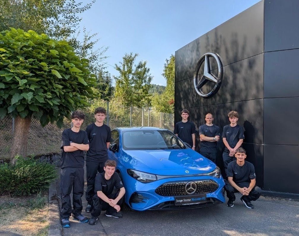 mehre junge Personen stehen neben einem blauen Mercedes, dahinter Gebüsch und eine schwarze Wand mit Mercedes Stern