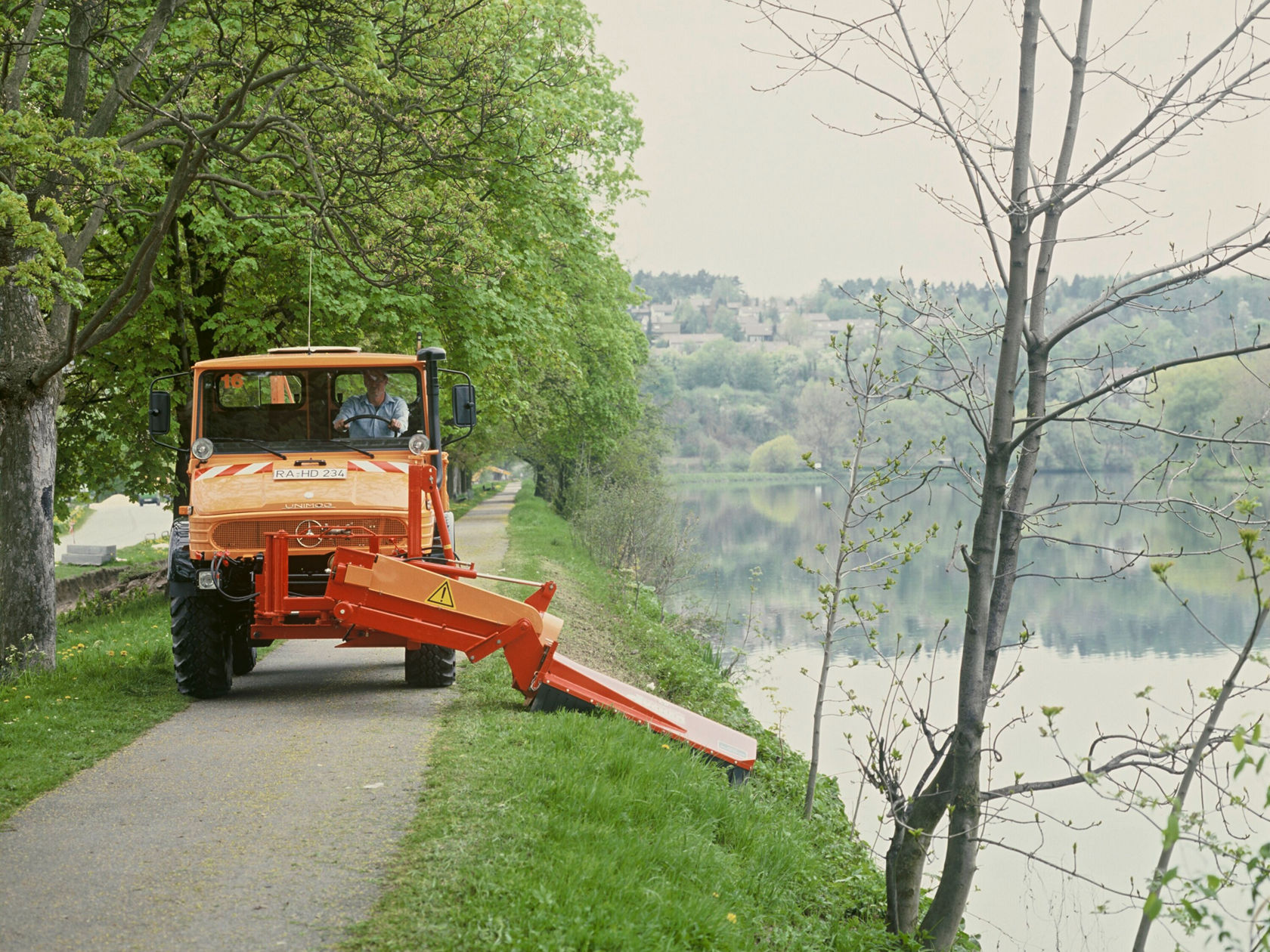 Een Mercedes-Benz Unimog met een maaier rijdt langs een weg aan het water, omgeven door bomen en groen, terwijl een persoon in de cabine zit.
