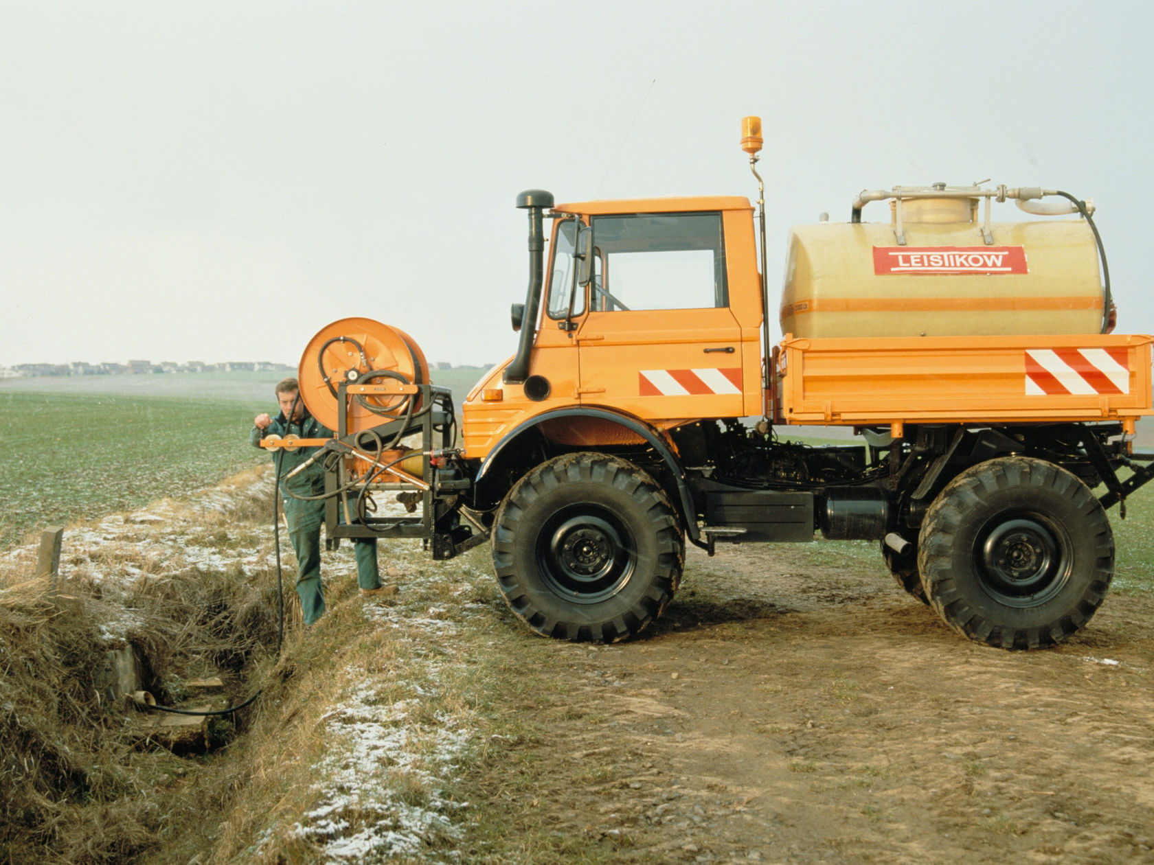 Een oranje Unimog van Mercedes-Benz staat naast een sloot, terwijl een arbeider een apparaat bedient dat met de Unimog verbonden is.