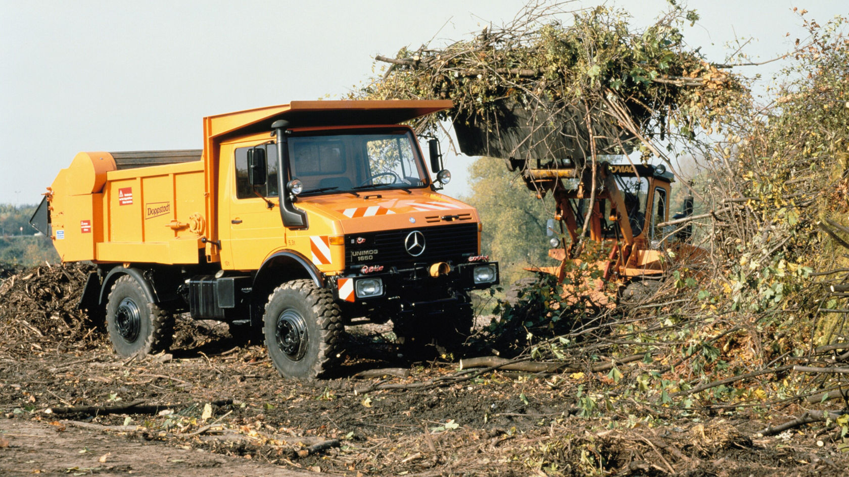 Een oranje Unimog van Mercedes-Benz staat in een ontbost gebied naast een houtharvester, omgeven door boomstammen en ongeploegde grond.