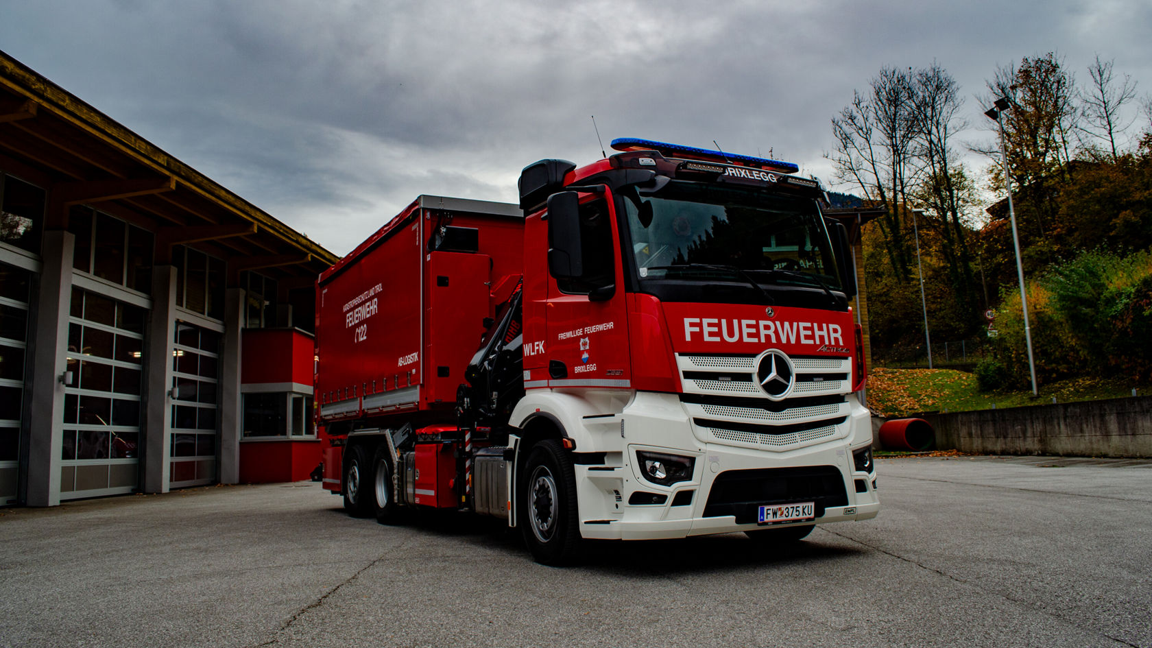A red Mercedes-Benz Actros in fire-fighting configuration stands in front of a fire station.