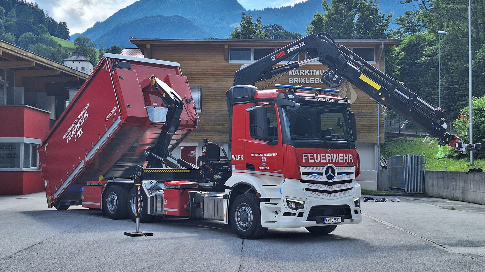 A red Actros fire-fighting vehicle with a container and a crane body is standing in front of a building. The container is being set down.