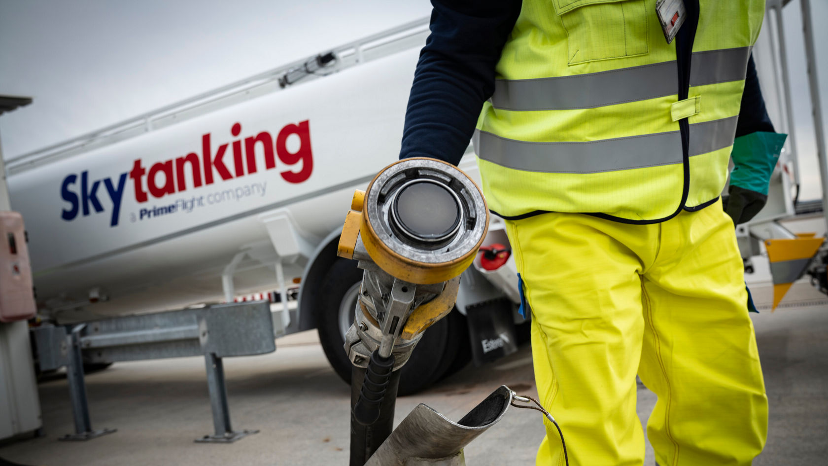 Ein Arbeiter in einer Sicherheitsweste und Regenhose hält einen Betankungsgriff. Im Hintergrund ist ein eEconic Tankwagen mit dem Logo „Skytanking“ zu sehen.