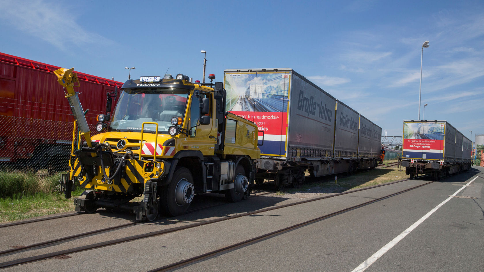 Ein gelber Mercedes-Benz Unimog steht neben Lastwagen auf einem Gleis. Der Unimog hat eine Anbauvorrichtung vorne und ist bereit für Einsätze im Transportwesen.