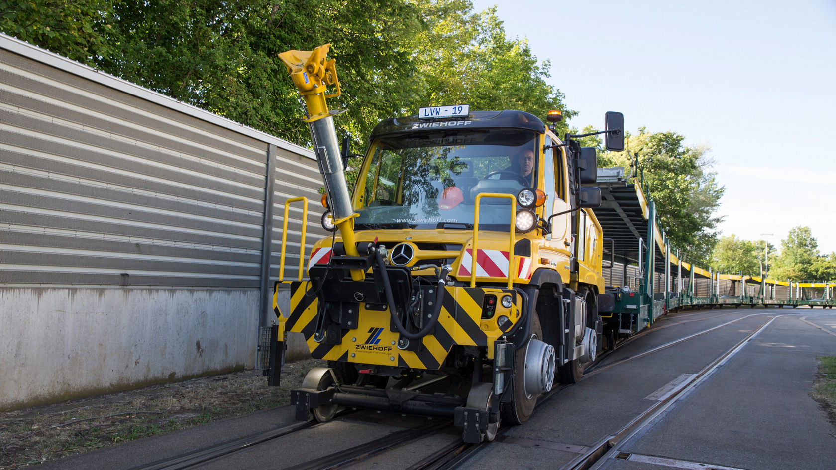 Gelber Mercedes-Benz Unimog mit einer Arbeitsvorrichtung steht auf Schienen neben einer grünen Straßenbahn. Der Unimog ist für Wartungsarbeiten an Gleisanlagen konzipiert.