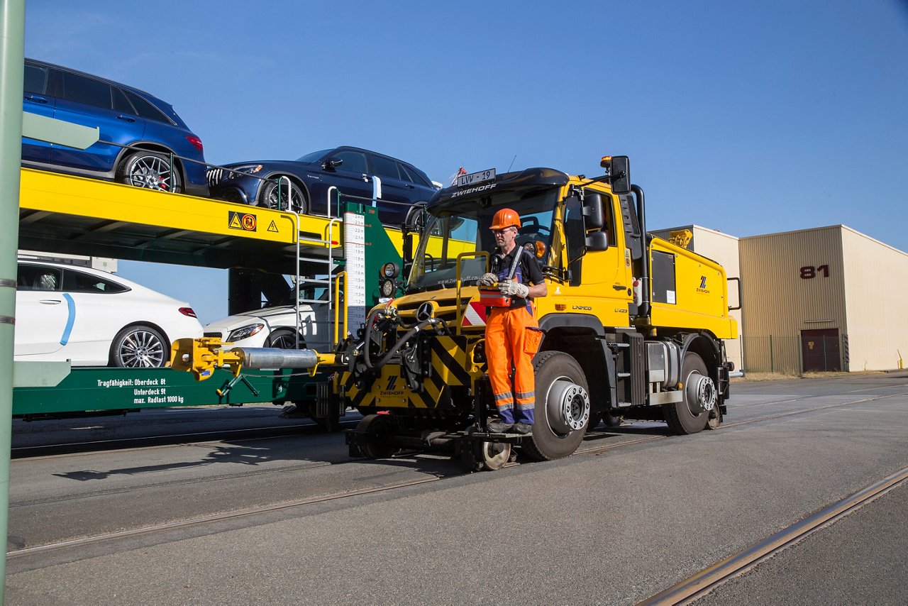 Ein Mercedes-Benz Unimog mit einem Arbeiter in orangefarbener Ausrüstung, der an einem Parkhaus steht, wo Fahrzeuge auf einem Autoanhänger transportiert werden.