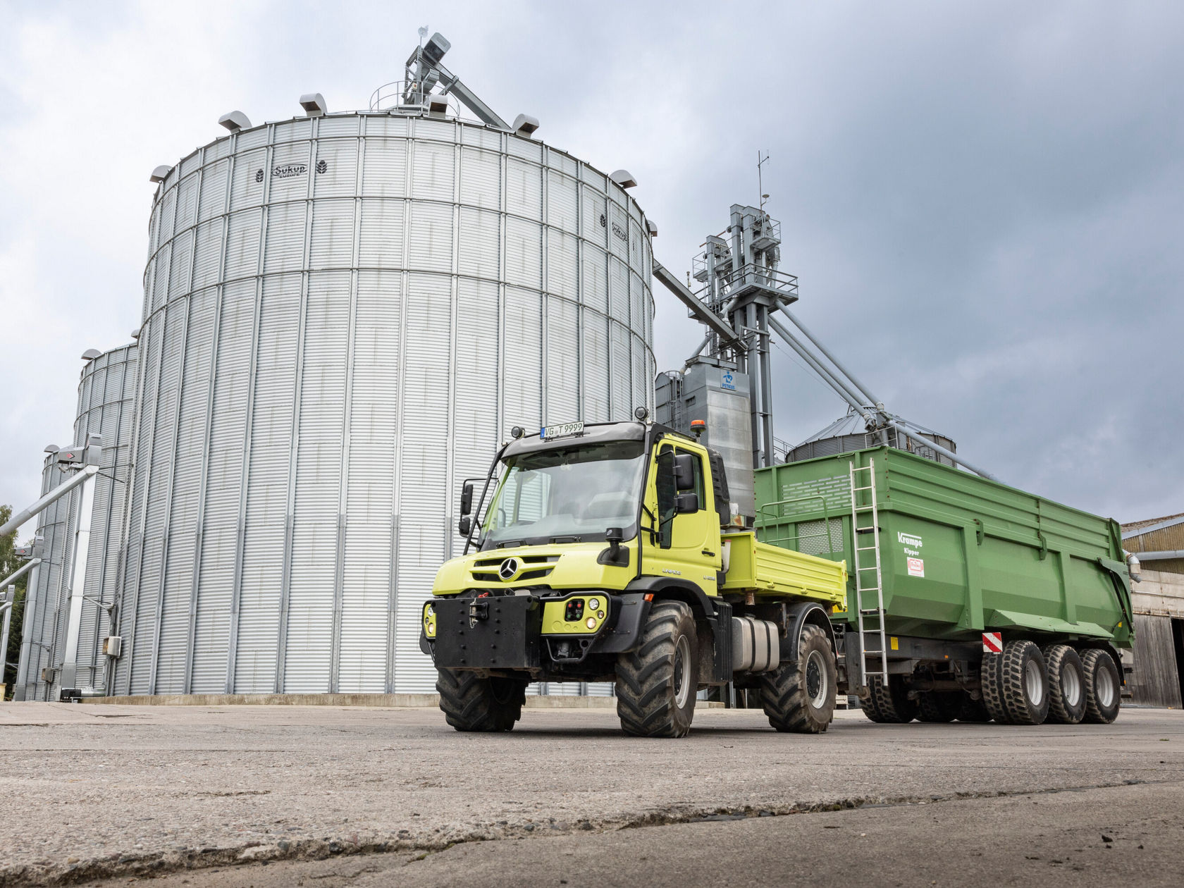 Un Unimog Mercedes‑Benz jaune se trouve devant une grande installation de silos avec une remorque verte. L'environnement a un caractère industriel sous un ciel gris.
