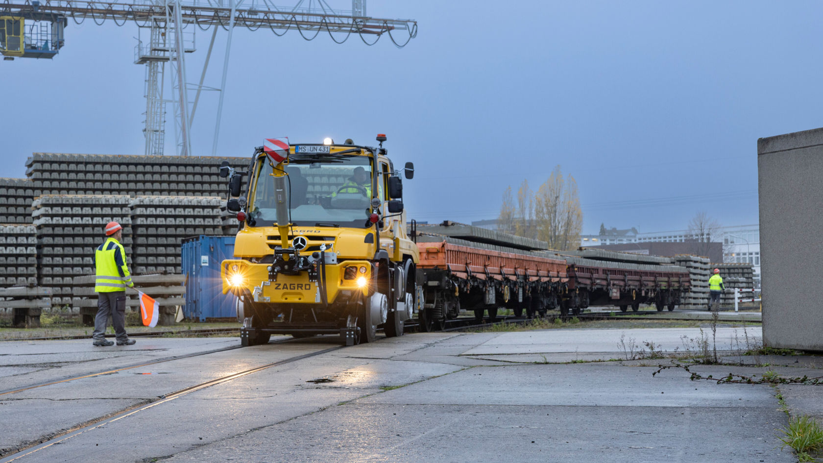 Ein gelber Zweiwege Unimog steht auf einem Gleis in einem Bahnbereich. Daneben steht eine Person in Warnkleidung mit einer Fahne.