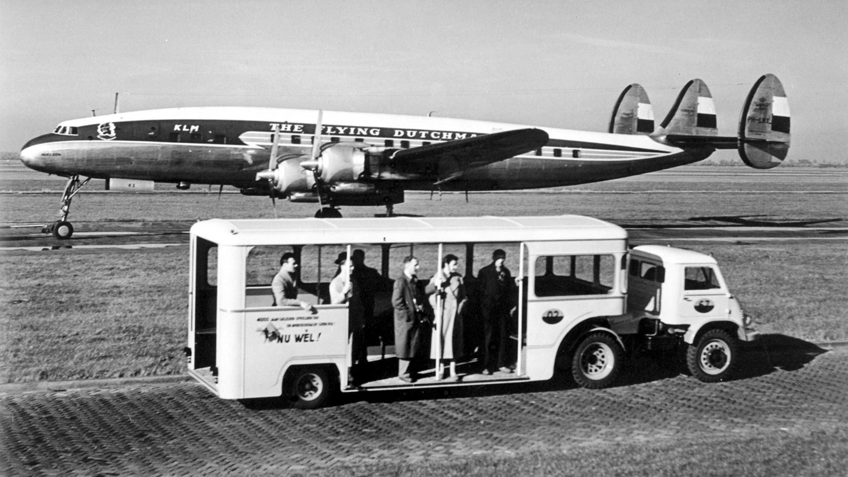 Eine Gruppe von Passagieren steigt aus einem historischen Bus vor einem KLM-Flugzeug, einem Lockheed Constellation, am Flughafen aus.