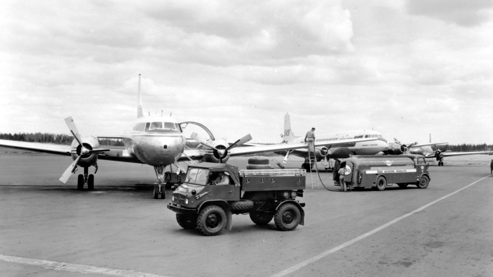 Ein Mercedes-Benz Unimog steht auf dem Flughafengelände, während ein Mitarbeiter mit einem Schlauch ein Flugzeug betankt. Im Hintergrund sind mehrere Flugzeuge zu sehen.