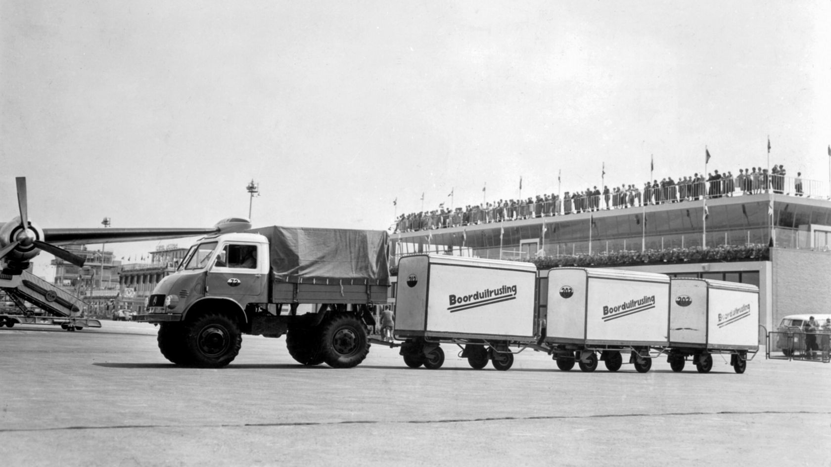 Ein Mercedes-Benz Unimog zieht mehrere Anhänger auf einem Flughafen, umgeben von Zuschauern auf einer Tribüne. Der Unimog ist für Transportaufgaben konzipiert.