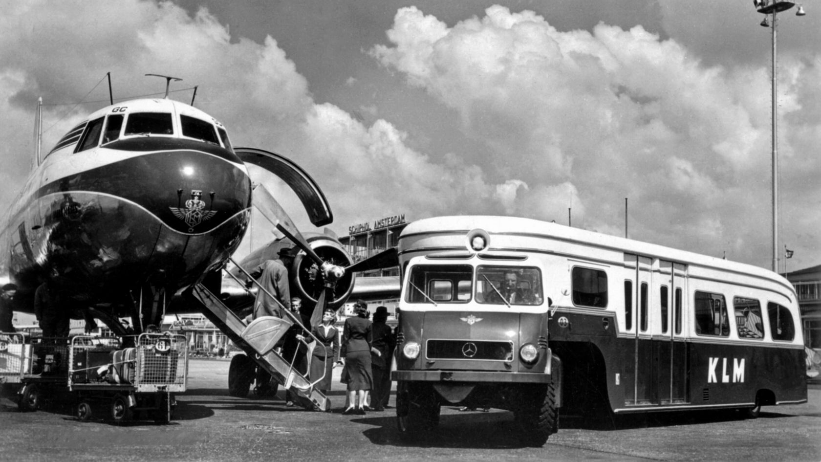 Ein Mercedes-Benz Unimog, der als KLM-Bus genutzt wird, steht neben einem Flugzeug am Flughafen, während Passagiere einsteigen.