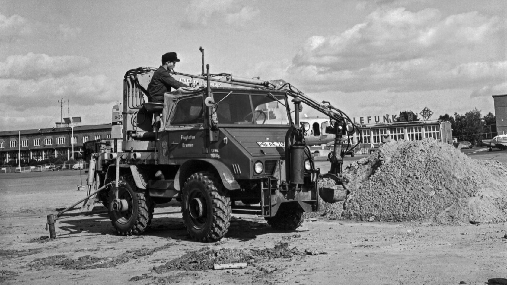 Ein Unimog von Mercedes-Benz, bedient von einem Mann, hebt Erde mit einer Geräteausrüstung auf einer Baustelle. Im Hintergrund ist ein Gebäude sichtbar.