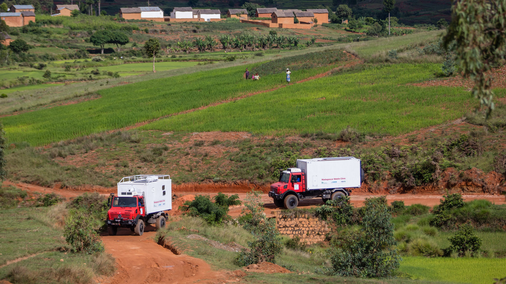 Deux véhicules Mercedes‑Benz robustes circulent sur un chemin non goudronné à travers un paysage rural verdoyant avec des champs et des arbres.