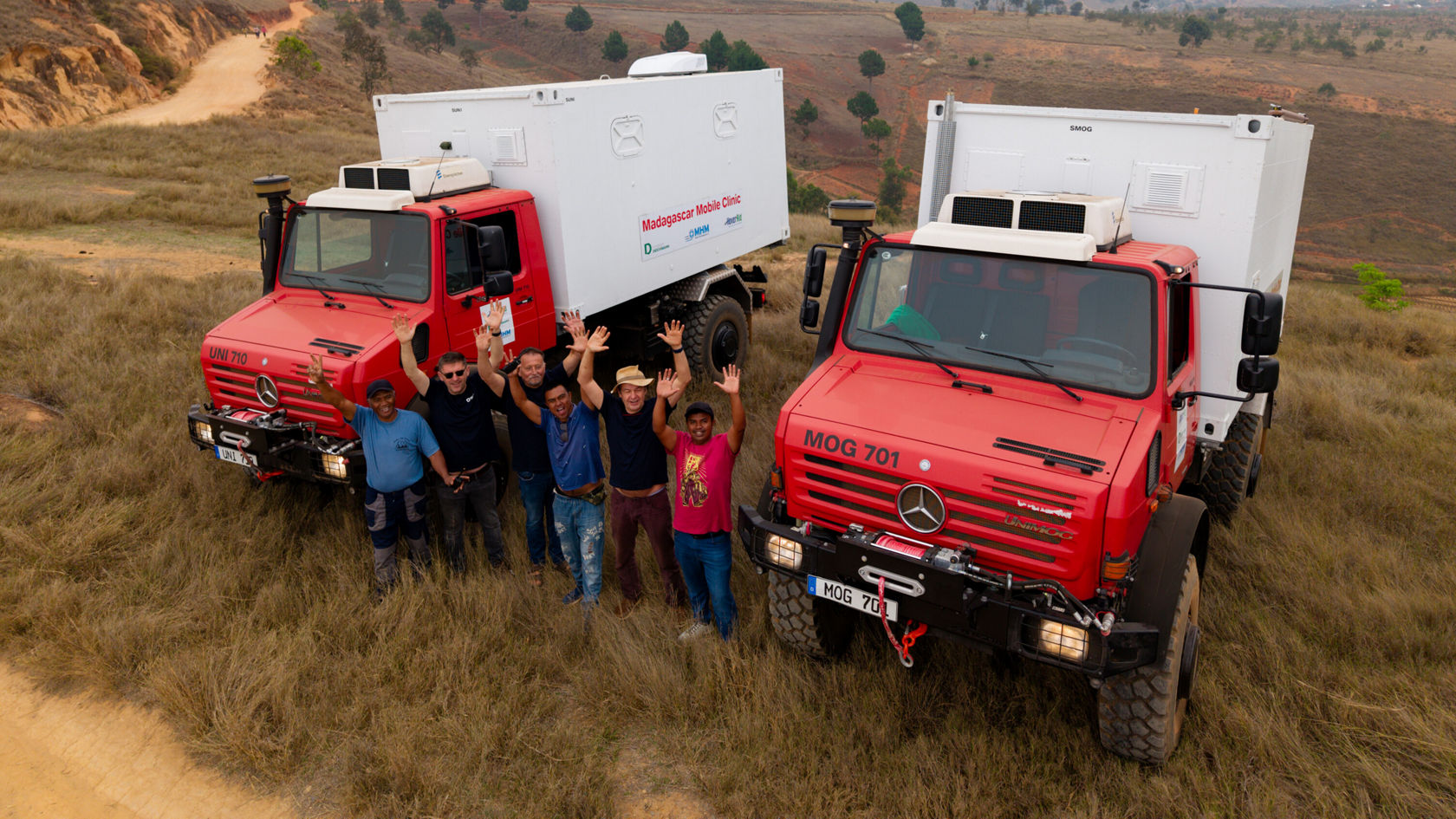 Deux Mercedes-Benz Trucks rouges se trouvent sur une prairie. Un groupe de sept personnes se tient devant les véhicules, souriant et faisant signe, entouré d'un paysage naturel.