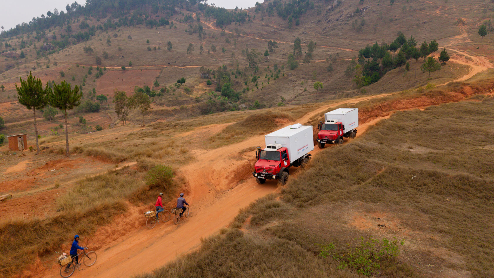 Des véhicules de livraison Mercedes‑Benz rouges roulent sur un chemin poussiéreux en milieu rural, tandis que des cyclistes se dirigent vers eux.